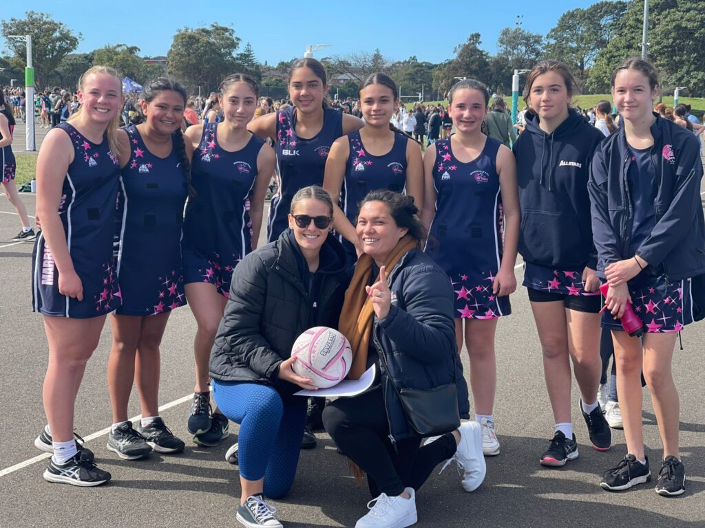 A group of young female netball players in dark blue and pink star-patterned uniforms posing together with two smiling coaches on an outdoor court.